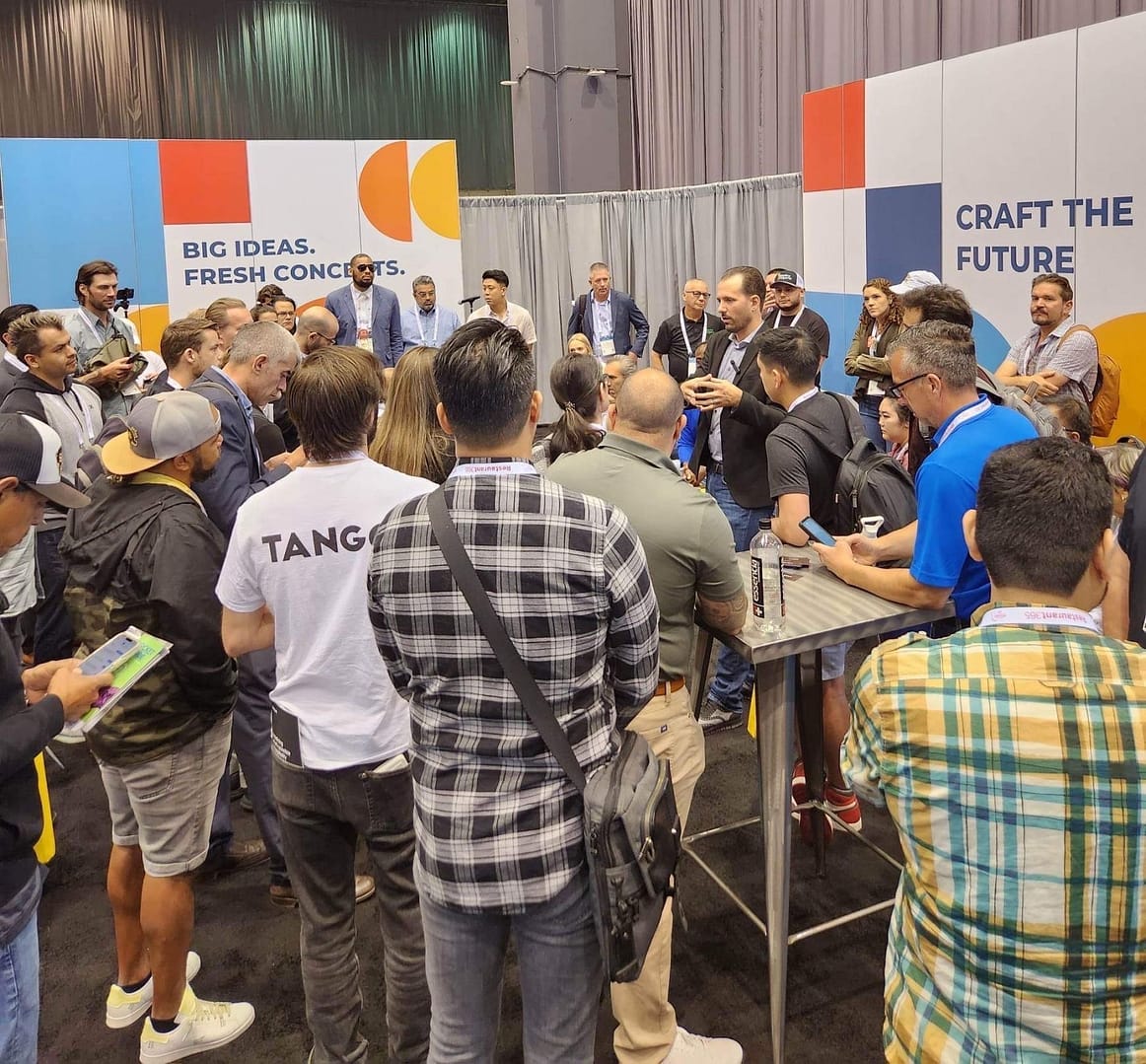 Trade-show crowd encircles a presenter speaking near booths that read “BIG IDEAS” and “CRAFT THE FUTURE.”