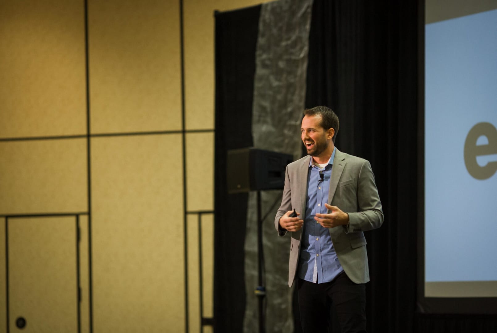 Presenter in a gray blazer and blue shirt laughs while speaking on a conference stage.
