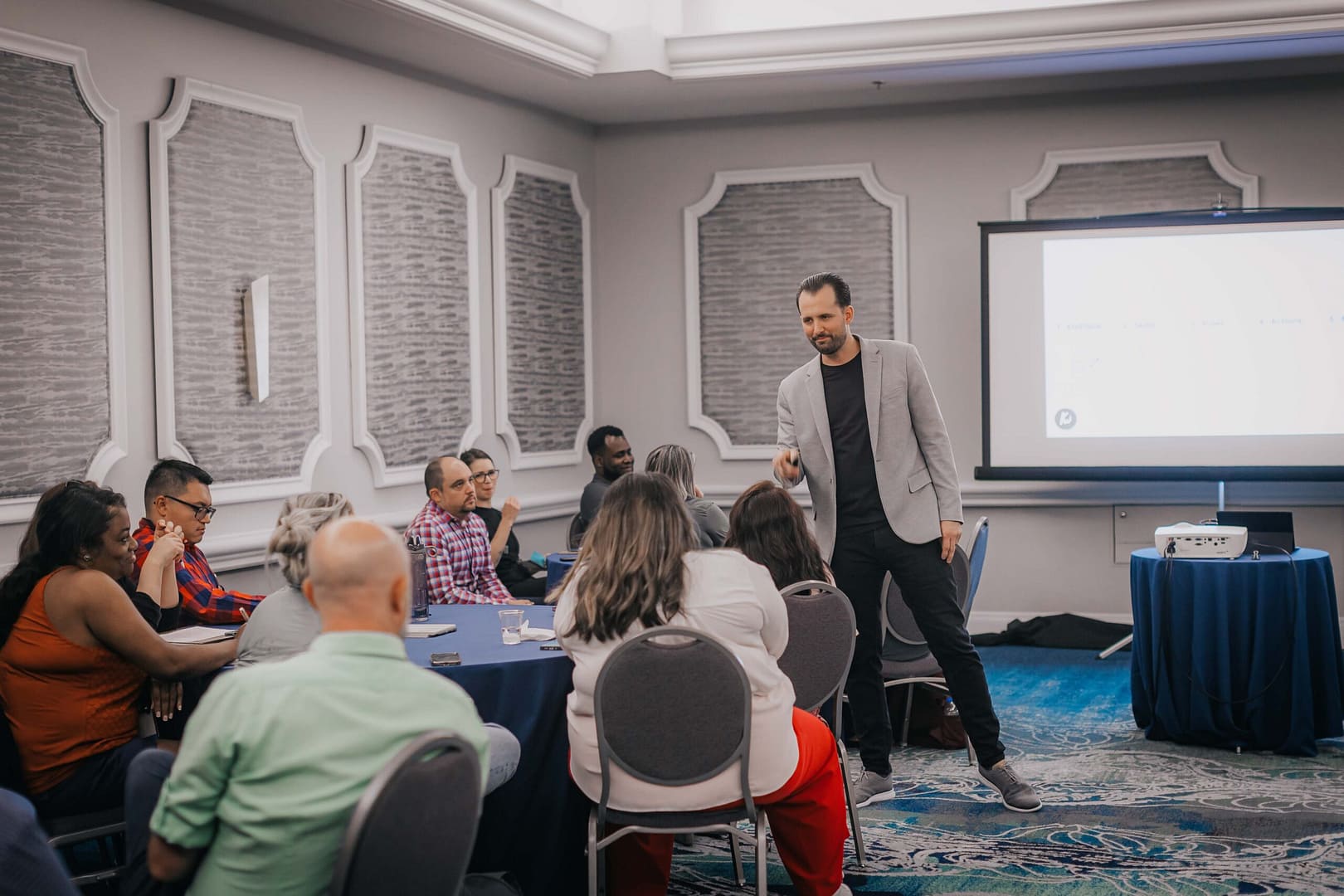 Workshop scene: a presenter in a gray blazer addresses attendees seated at blue-covered tables.