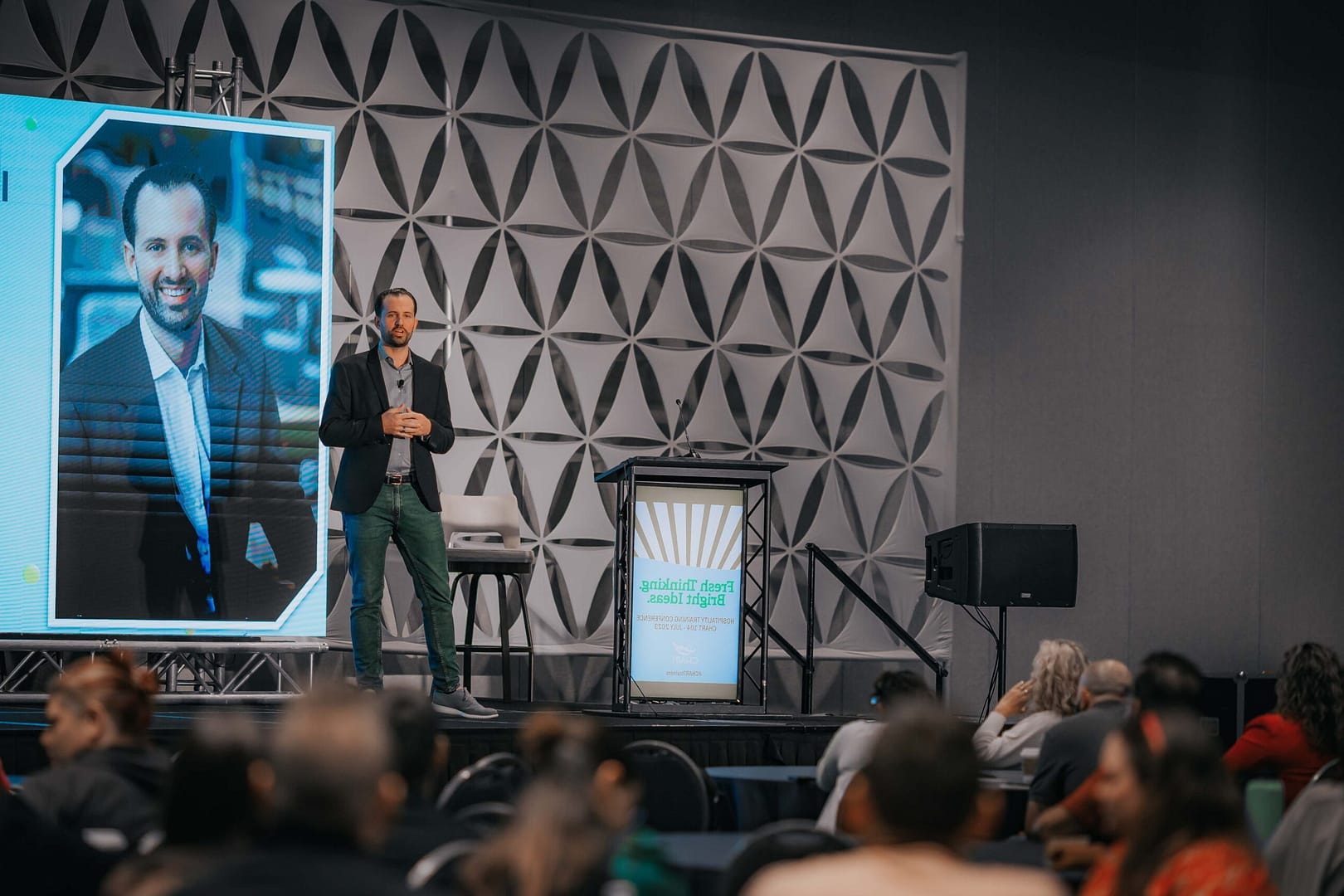 Keynote speaker in a blazer stands on a large stage beside a tall screen showing his portrait.