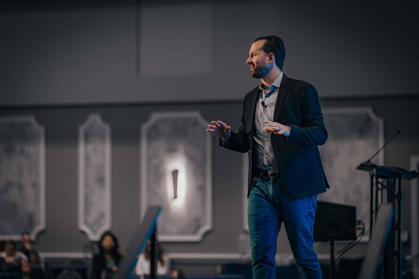 Full-body view of a presenter in a dark blazer and jeans speaking on a wide conference stage.