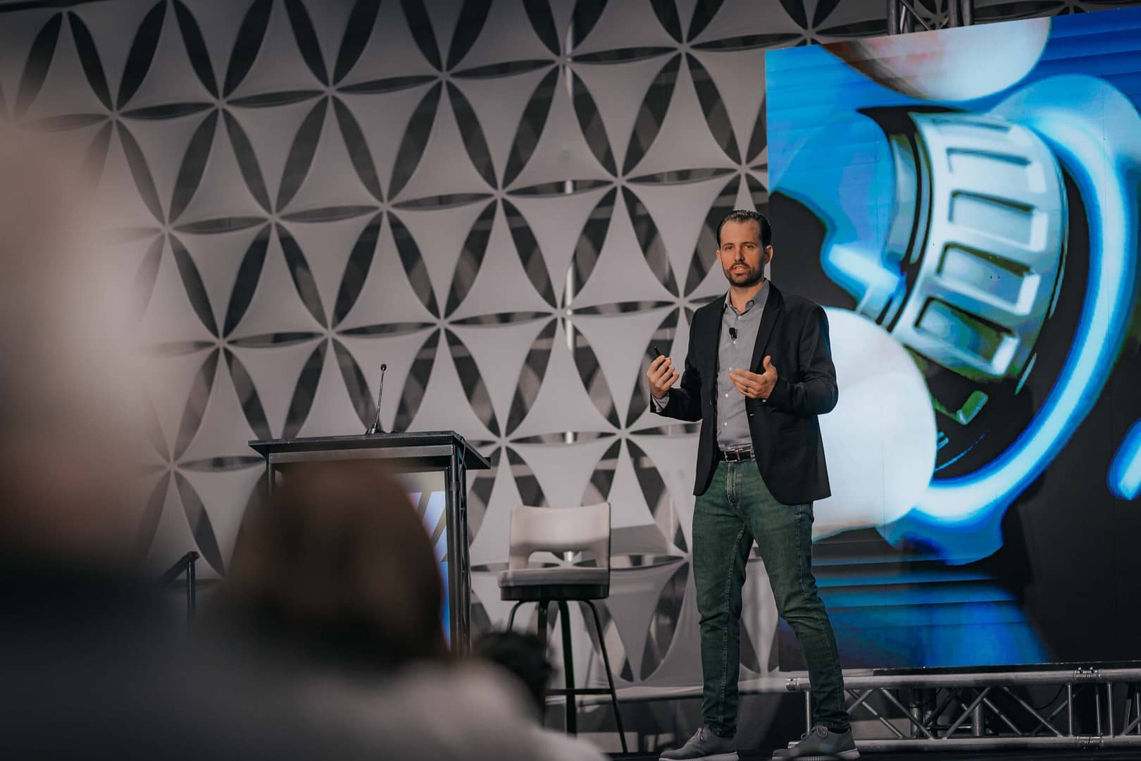 Keynote speaker in a tan suit with a headset mic emphasizes a point on a geometric stage.