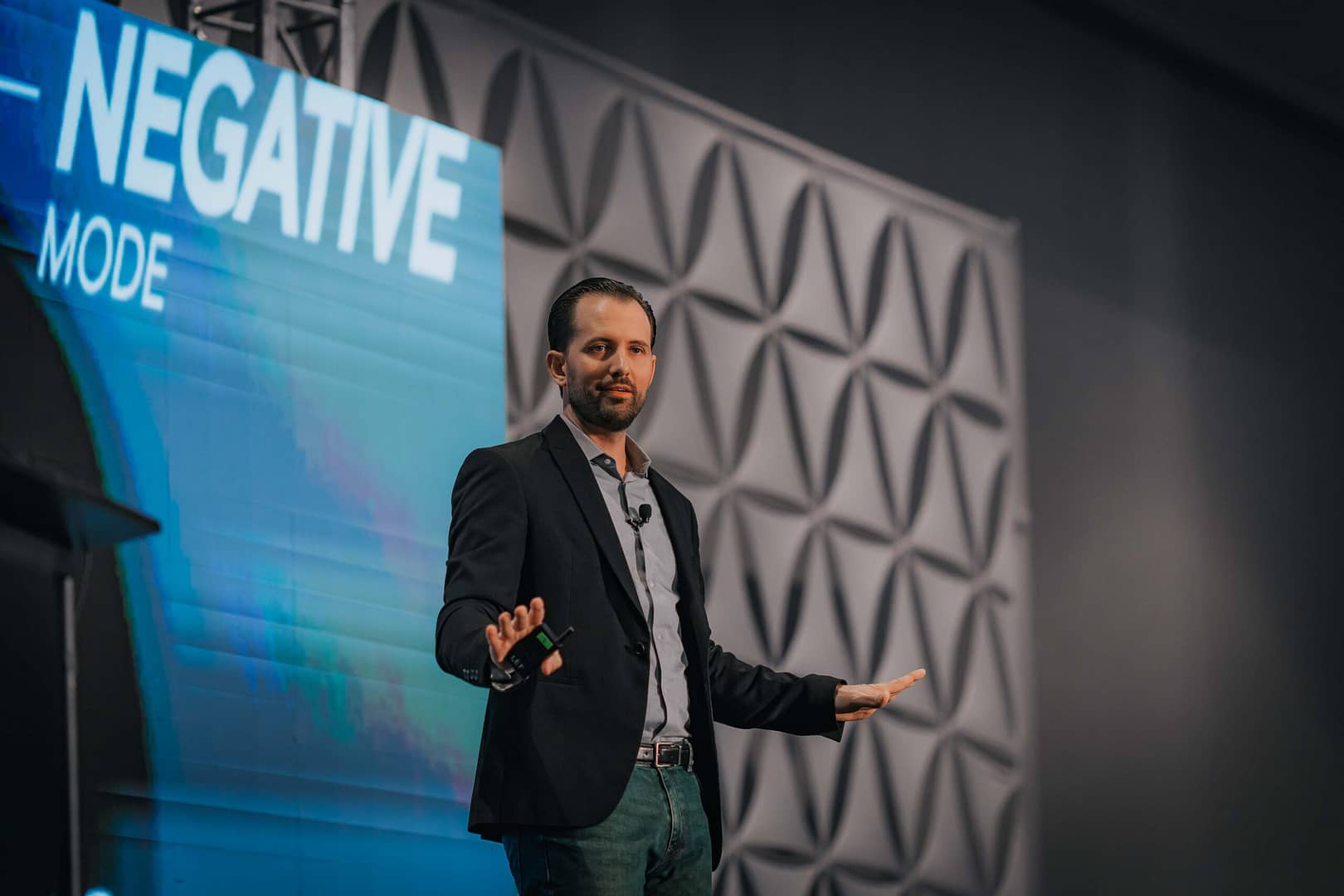Speaker in a dark blazer stands beside a screen titled “NEGATIVE MODE,” hands extended.