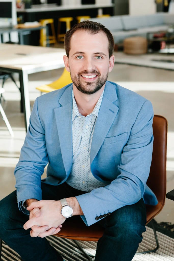 Professional headshot of a smiling man seated in a modern office, wearing a light-blue blazer and open-collar shirt.