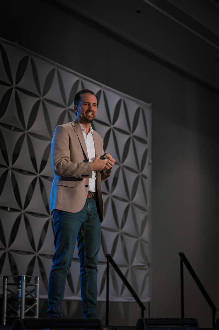 Full-body view of a presenter in a tan blazer and jeans addressing the audience from an elevated stage.