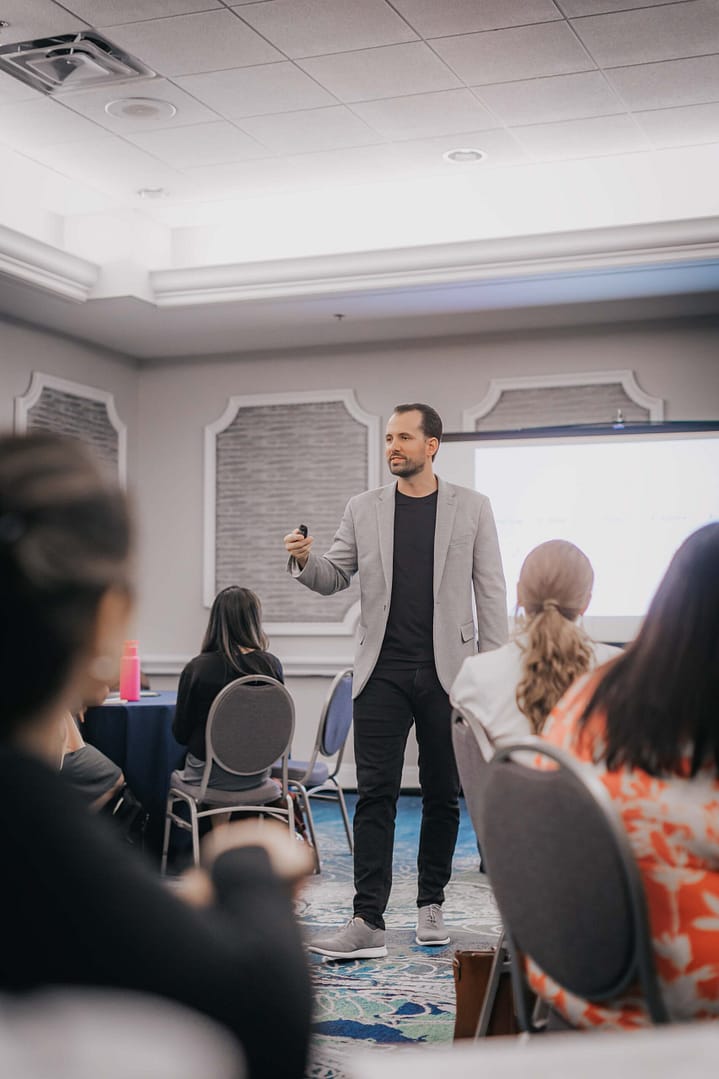 Presenter in a light-gray blazer engages attendees at the front of a workshop setting.