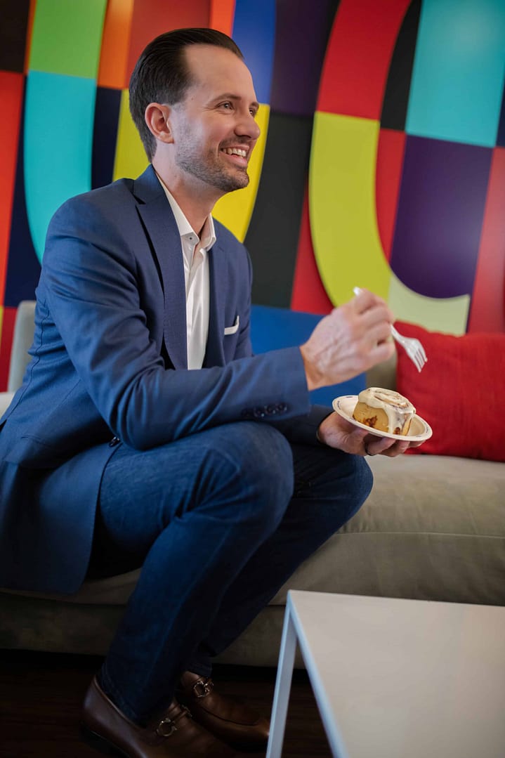 Smiling man in a navy suit sits on a couch eating a cinnamon roll against a vibrant multicolored wall.